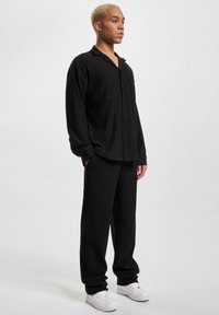 Cămașă neagră cu nasturi, cu textură verticală și canelată, cu două buzunare la piept, asortată cu pantaloni negri largi și adidași albi.