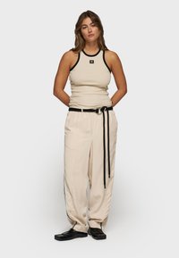 Top sin mangas de canalé beige con ribetes negros, combinado con pantalones crema de corte holgado que presentan un cinturón negro y franjas laterales. Zapatos negros brillantes.