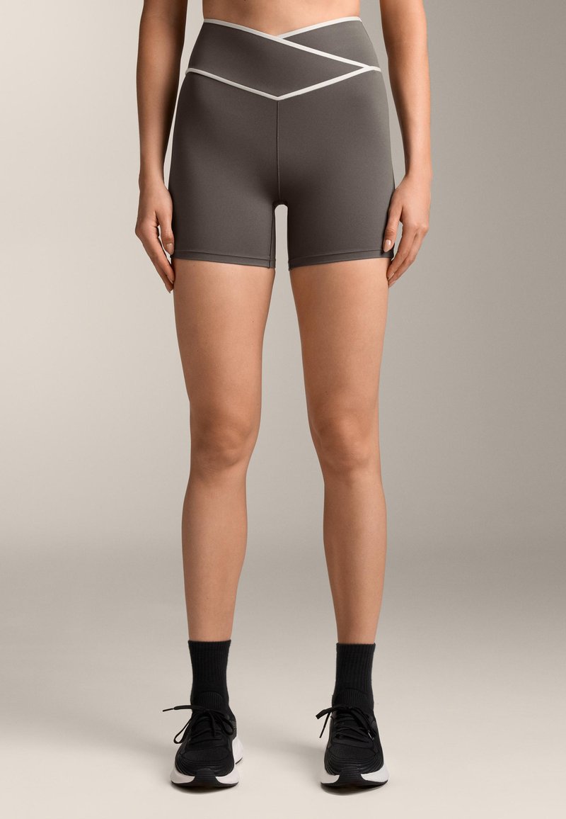 Sive atletske kratke hlače z visokim, prešitvenim pasom. Gladka tekstura, dolžina do sredine stegna, v kombinaciji s črnimi športnimi copati in kratkimi nogavicami.