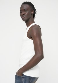 Λευκό αμάνικο τοπ σε εφαρμοστή γραμμή, κατασκευασμένο από ριμπ ύφασμα. Το μοντέλο στέκεται πλαγίως, φοράει μπλε τζιν και έχει το ένα χέρι στην τσέπη.