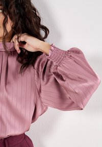 Hedvábná, růžová blůza s vertikálními pruhy, která má nařasené rukávy a elastické manžety. Hladká textura a splývavý design zdůrazňují eleganci.