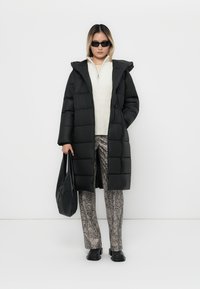 Lang sort fyldig frakke med hætte, parret med en cremefarvet sweater og mønstrede grå slangeprintbukser. Accessories inkluderer sorte solbriller og en taske.