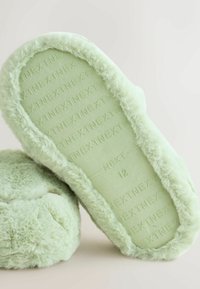 Pantuflas verdes peludas con un exterior de felpa y un interior suave. La suela tiene texto en relieve y una etiqueta de tamaño, con una forma redondeada.