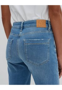 Bakvy av en person som bär ljusblå denimjeans med två bakfickor och en brun läderlogotyp på midjan.