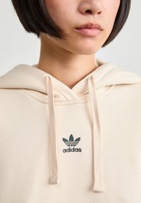 Sweatshirt bege feito de um tecido suave, com bolso canguru, cordões e um logótipo da Adidas em verde escuro cosido no peito.