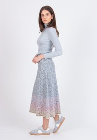 Kvinde i lyseblå ribbet turtleneck, mønstret midi-nederdel med blå og lyserød gradient, og lyseblå sneakers, der står sidelæns på hvid baggrund.