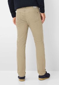 Beige Baumwoll-Chinos mit schlankem Schnitt, ausgestattet mit zwei Gesäßtaschen und einem schlichten Bundlabel. Getragen mit dunkelblauen Schuhen.