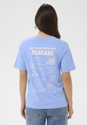 Vrouw met lang donker haar, gezien van achteren, draagt een lichtblauw T-shirt met een wit pannenkoekenrecept en illustraties op de achterkant.