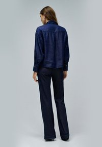 Camisa de mezclilla azul marina con mangas largas, puños abotonados y un cuello clásico; combinada con jeans oscuros acampanados, ambos con una textura suave.