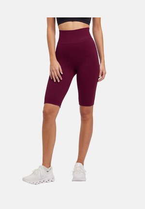 Frau trägt hoch taillierte, strukturierte dunkelrote Biker-Shorts und weiße Laufschuhe, steht mit entspannten Händen seitlich am Körper.