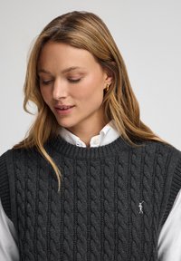 Kolgrå stickad väst med en texturerad kabelmönster, rund halsringning och ribbad ärmhål, bärs över en vit knäppskjorta.