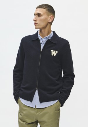 Mand med kort hår iført en sort lynlåsstriktrøje med et hvidt "W"-logo, en blåstribet skjorte og beige bukser, ser til venstre med hænderne i lommerne.