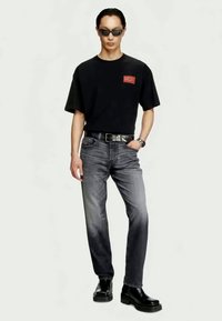 T-shirt preta com o logótipo vermelho "DIESEL", combinada com jeans cinzentos e botas de cano curto em pele pretas. Os acessórios incluem óculos de sol e um relógio.