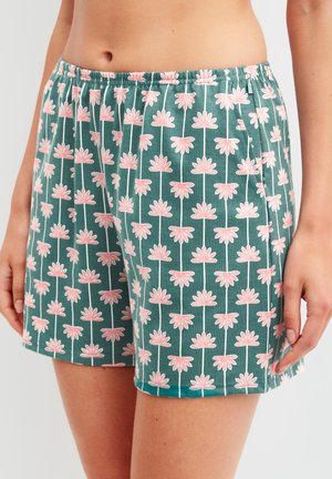 Taille tot halverwege de dij van een persoon die turquoise korte broek draagt met een verticale bloemenpatroon in roze en wit, een elastische tailleband en zijzakken.
