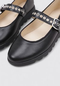 Zapatos mary-jane de cuero negro con punta redondeada, una correa texturizada con ojales de plata y una suela de goma gruesa.