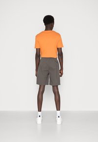 T-shirt en coton orange, vue de dos, associé à un short gris. Modèle portant des baskets blanches avec des accents noirs et des chaussettes blanches. Design simple.