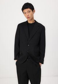 Blazer noir ajusté avec deux boutons, col à revers et poches avant. Porté par-dessus un top noir, présentant un tissu lisse et un design structuré.