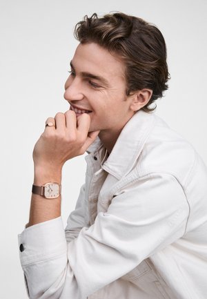 Montre carrée en or rose avec un cadran beige et un bracelet en cuir marron, portée sur une manche en denim clair, présentant de subtils détails de coutures.