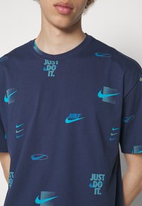 Mladý člověk v námořnickém modrém tričku s různými modrými logy Nike a slogany "Just Do It", stojící na jednoduchém pozadí.