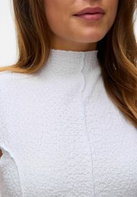 Witte textuur top met een hoge halslijn, voorzien van een subtiel patroon en een sluiting aan de voorkant, gezien vanaf de borst omhoog tegen een neutrale achtergrond.