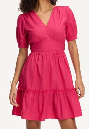 Femme portant une robe courte rose vif à manches courtes avec un col en V, une taille cintrée, une jupe froncée et un détail volanté près de l'ourlet.