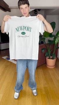 Camiseta de algodón blanca con "NEWPORT 1957 Sporting Goods" impreso en verde. Corte holgado, mangas cortas, sostenida por una persona que lleva unos pantalones de mezclilla y zapatillas plateadas.