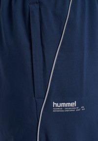 Mörkblå techwear-shorts i strukturerad tyg, grå kant längs sidan och en framträdande vit logotyp nära fållen.