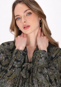 Bluză verde cu model, cu mâneci lungi și puffate și detaliu cu fundă la gât. Prezintă nasturi decorativi pe manșete.