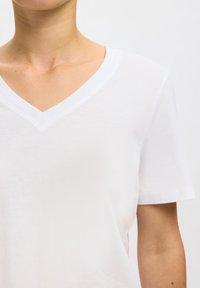 Oberkörper einer Person, die ein schlichtes weißes T-Shirt mit V-Ausschnitt und kurzen Ärmeln vor neutralem Hintergrund trägt.