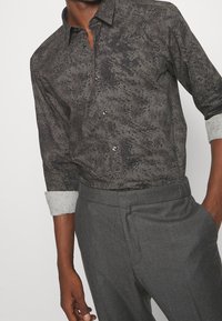 Chemise à motifs sombres avec une texture lisse, dotée d'un col rabattable et d'une patte de boutonnage. Manches avec poignets en tissu gris plus clair.