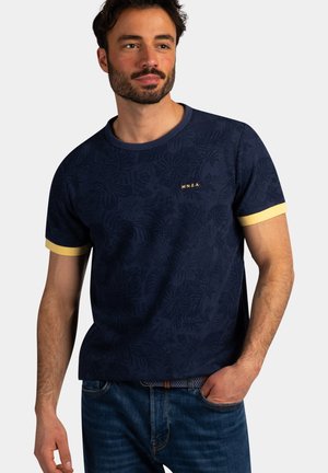 Man draagt een marineblauw T-shirt met bloemenpatroon, gele mouwboorden en een klein "N.Z.A."-logo, gecombineerd met een blauwe spijkerbroek en hand in de zak.