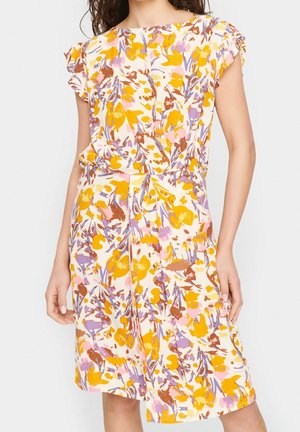 Femme portant une robe mi-longue avec un motif floral coloré en jaune, rose, marron et violet sur fond blanc.