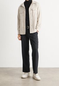 Chaqueta beige abotonada con dos bolsillos en el pecho, cuello de tortuga negro, pantalones oscuros y zapatillas blancas con acentos beige. Textura suave.