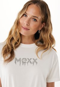 Bílý bavlněný tričko s grafickým designem, na kterém jsou kovové stříbrné korálky tvořící nápis "MXX" ve stylu kapající barvy. Krátké rukávy, kulatý výstřih.