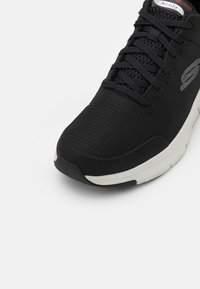 Chaussure de sport noire avec une tige en mesh texturé, dotée de lacets ton sur ton et d'une semelle intérieure ArchFit. Semelle intermédiaire blanche avec une légère courbe.