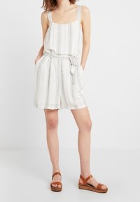 Vit randig jumpsuit med bälte i midjan, fyrkantig halsringning och dekorativa detaljer. Tillsammans med bruna sandaler med träsål.