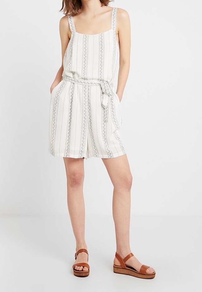 Vit randig jumpsuit med bälte i midjan, fyrkantig halsringning och dekorativa detaljer. Tillsammans med bruna sandaler med träsål.