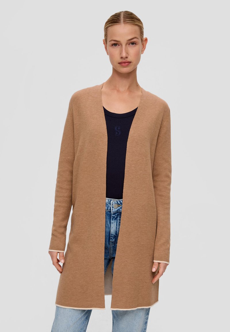Cardigan long léger couleur brun clair en matière douce, avec un design ouvert à l'avant, rehaussé d'une bordure crème contrastante au niveau des poignets et de l'ourlet. Porté par-dessus un haut sombre.