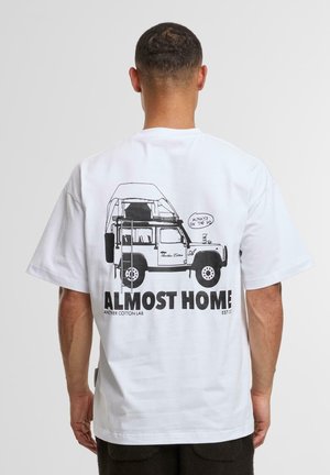 White cotton t-shirt featuring a black graphic illustration of a vehicle with a rooftop tent. Text below reads "ALMOST HOME" and "ANOTHER COTTON LAB."