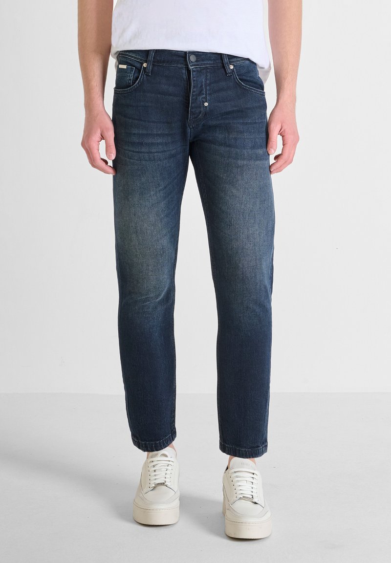 Jeans de mezclilla azul oscuro con un ajuste slim, piernas estrechas y un ligero desgaste. Cuenta con cinco bolsillos y un cierre de botón. Combinados con zapatillas blancas.