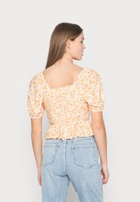 Blusa floral com pregas, mangas bufantes, padrão laranja e branco, decote quadrado nas costas e bainha peplum. Usada com jeans azul claro.