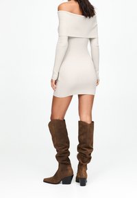 Robe en tricot beige à épaules dénudées avec manches longues, silhouette ajustée. Associée à de hautes bottes en daim marron, souples, avec talons épais.