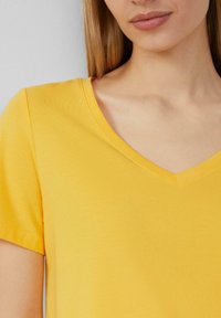 Gelbes Baumwoll-T-Shirt mit V-Ausschnitt und kurzen Ärmeln. Glatte Textur, taillierte Passform und minimale Nähdetails am Ausschnitt.