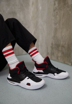 Person trägt schwarze, weiße und rote Basketballschuhe mit weiß-roten gestreiften Socken und sitzt an einer Betonwand auf einem hellgrauen Boden.