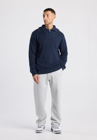 Man standing with hands in pockets wearing a navy blue zip sweater, light gray sweatpants, and white sneakers on a plain white background.
