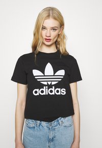 Must värvi puuvillane T-särk, millel on suur valge Adidas'i logo ja kolme triipu stiliseeritud nelilehe märgi all. Lühikesed varrukad, sportlik lõige.