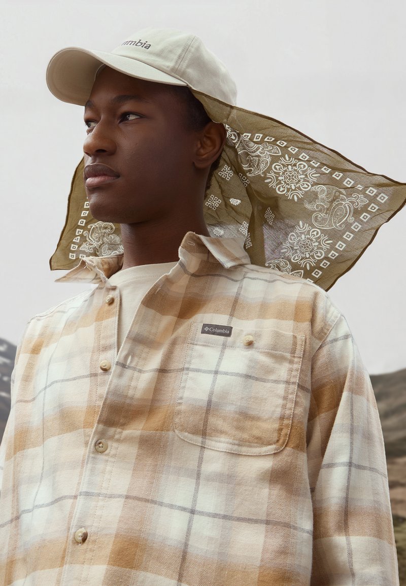 Camisa de cuadros beige y marrón con un bolsillo en el pecho, camiseta de color claro debajo y gorra beige con un pañuelo estampado atado por detrás.