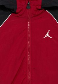 Κόκκινο αθλητικό μπουφάν με φερμουάρ πλήρους μήκους, μαύρες λεπτομέρειες, κουκούλα με επένδυση από δίχτυ και λευκό λογότυπο Jumpman στο μπροστινό μέρος.
