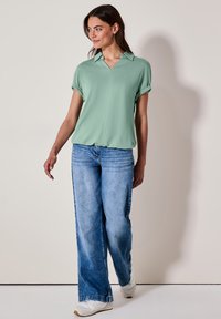 Hellgrünes Kurzarmoberteil mit Kragen und Reißverschlussfront, kombiniert mit blauen weit geschnittenen Jeans und weißen Sneakern auf einem neutralen Hintergrund.