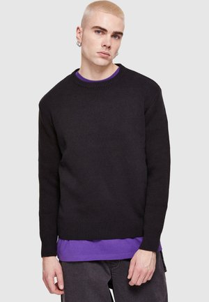 Schwarzer Strickpullover mit Rundhalsausschnitt und gerippten Bündchen. Über einem lila T-Shirt getragen, das eine lockere Passform und eine strukturierte Textur aufweist.